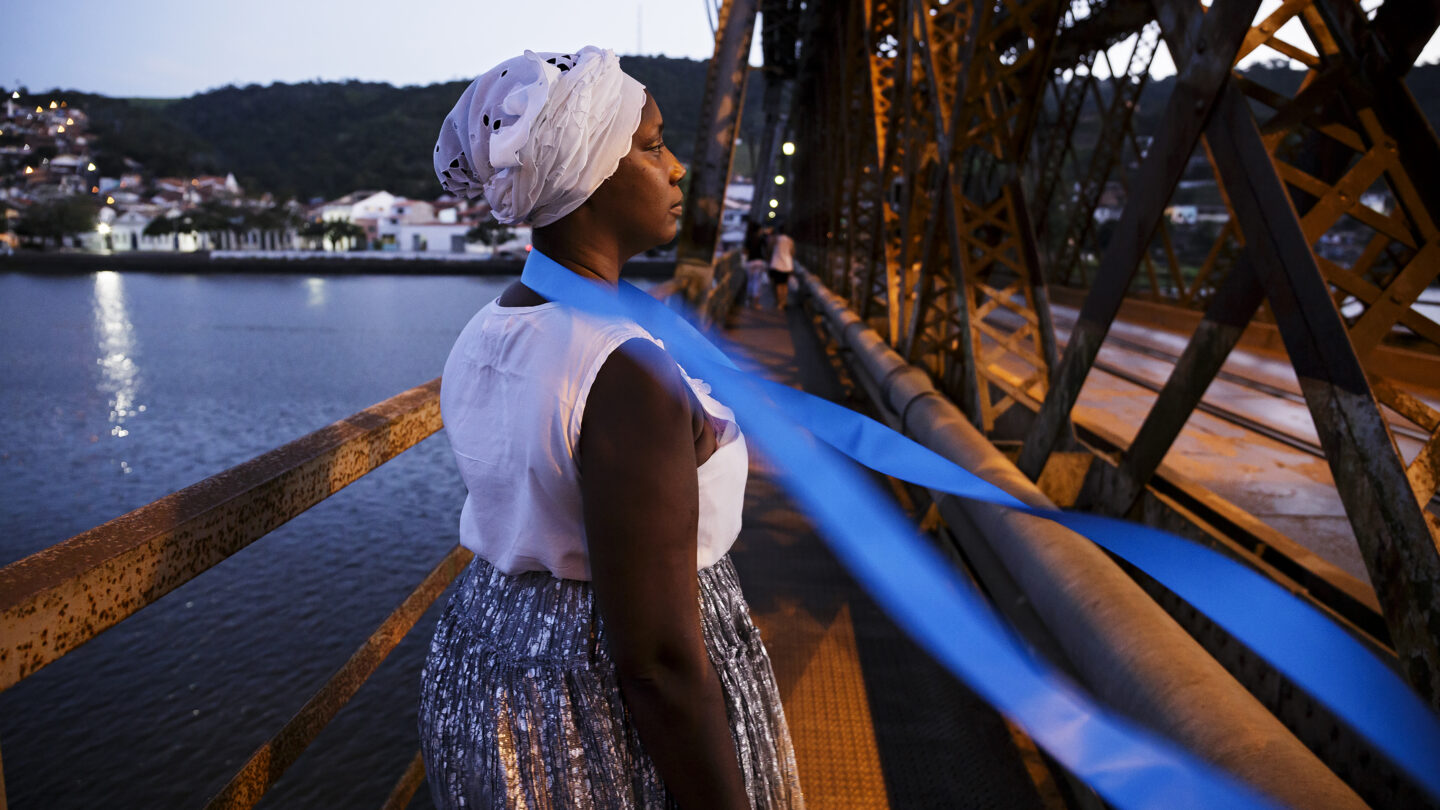 Fotografia de uma mulher negra de perfil olhando para o lado direito. Ela usa roupas brancas e um lenço azul no pescoço que é movimentado pelo vento, formando uma diagonal na imagem. A mulher está em cima de uma ponte.