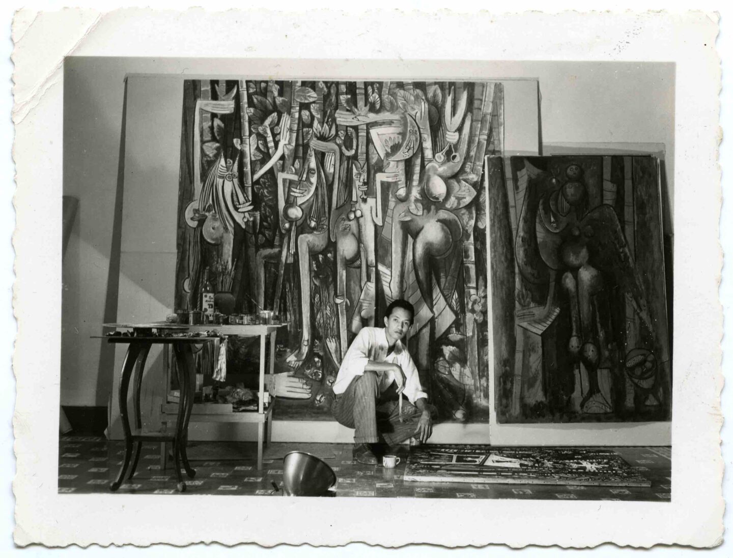 Foto antiga em preto e branco do artista diante de duas telas em grande formato, ele está agachado diante de uma outra tela que está no chão, dentro de seu estúdio