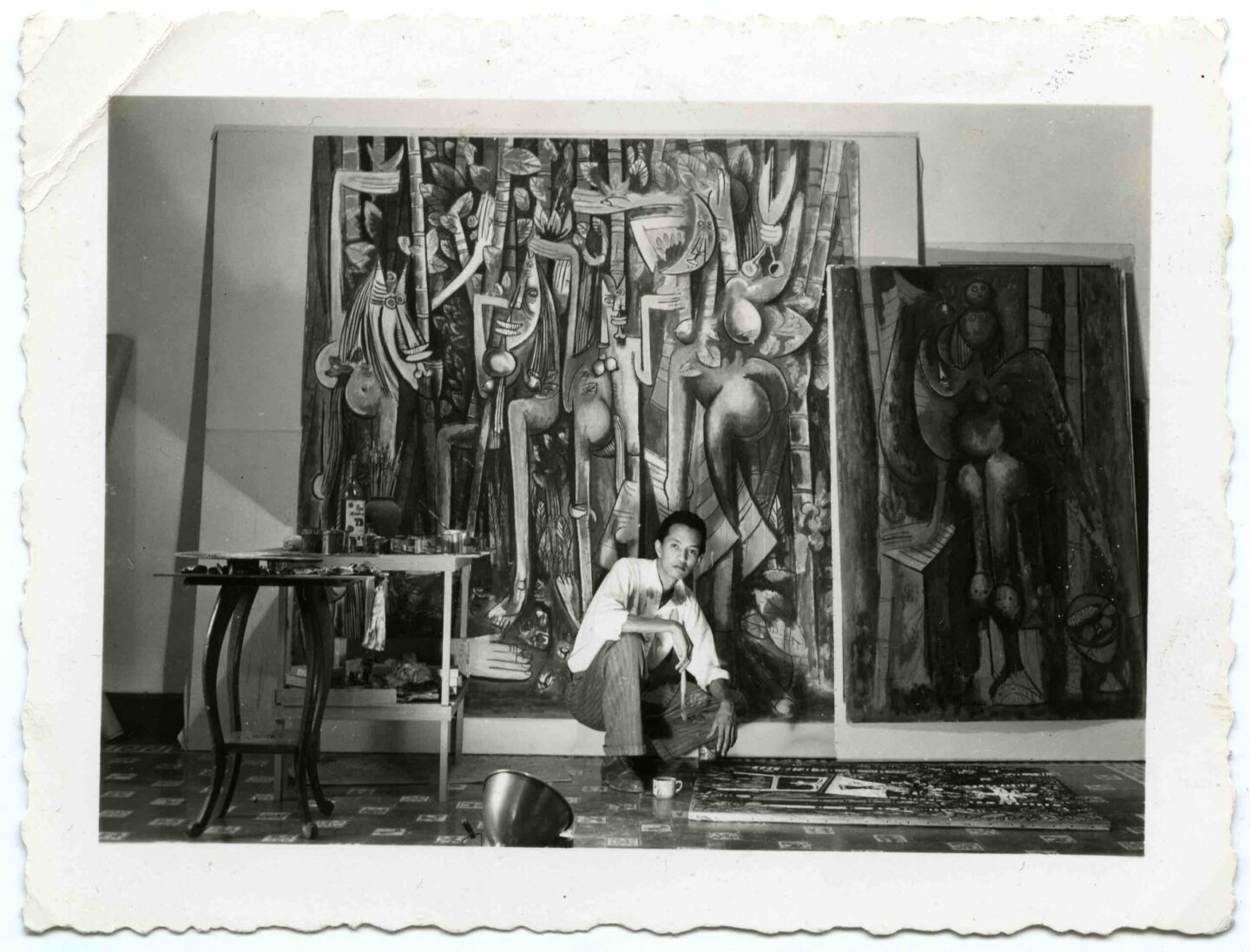 Foto antiga em preto e branco do artista diante de duas telas em grande formato, ele está agachado diante de uma outra tela que está no chão, dentro de seu estúdio