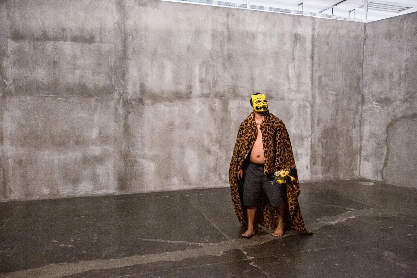 Foto de performance de Denilson Baniwa caminhando pelo Pavilhão da Bienal durante a 33ª Bienal, diante de uma parede cinza e chão preto. O artista usa uma máscara de onça, casaco longo com estampa de onça e bermuda, e caminha descalço.