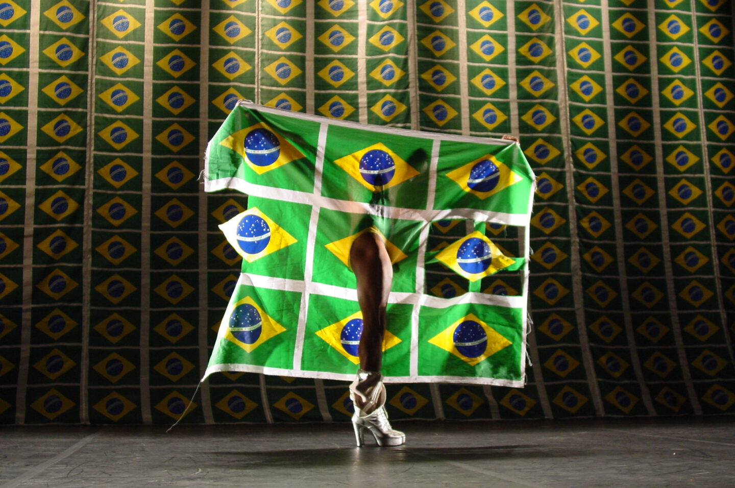 Vista de performance de Luiz de Abreu que mostra uma parede de bandeiras do Brasil em série ao fundo e uma pessoa rasgando um pano com bandeiras do Brasil em série com sua perna, usando um salto alto prateado.