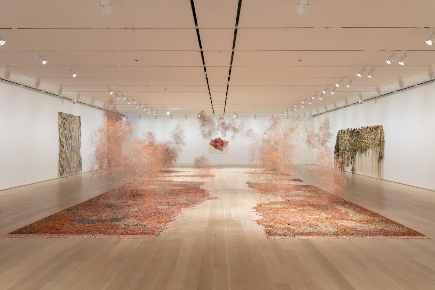 Vista de instalação de Igshaan Adams no The Art Institute of Chicago. A instalação é composta de duas partes que ficam no chão, abrindo um caminho vazio entre elas, e "nuvens" de poeira que são feitas de miçangas vermelhas, amarelas e laranjas, assim como o chão.