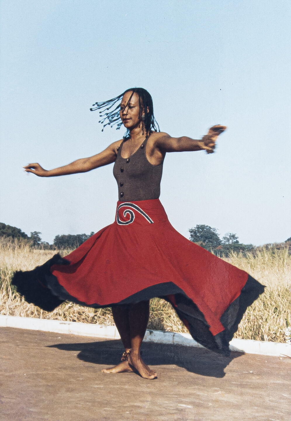 Foto de Inaicyra Falcão realizando performance. A artista é uma mulher negra e está rodando com os braços estendidos, vestindo uma regata marrom e uma saia vermelha com barra preta que está esvoaçando com o rodar. Ela está numa estrada de concreto em frente a um campo.