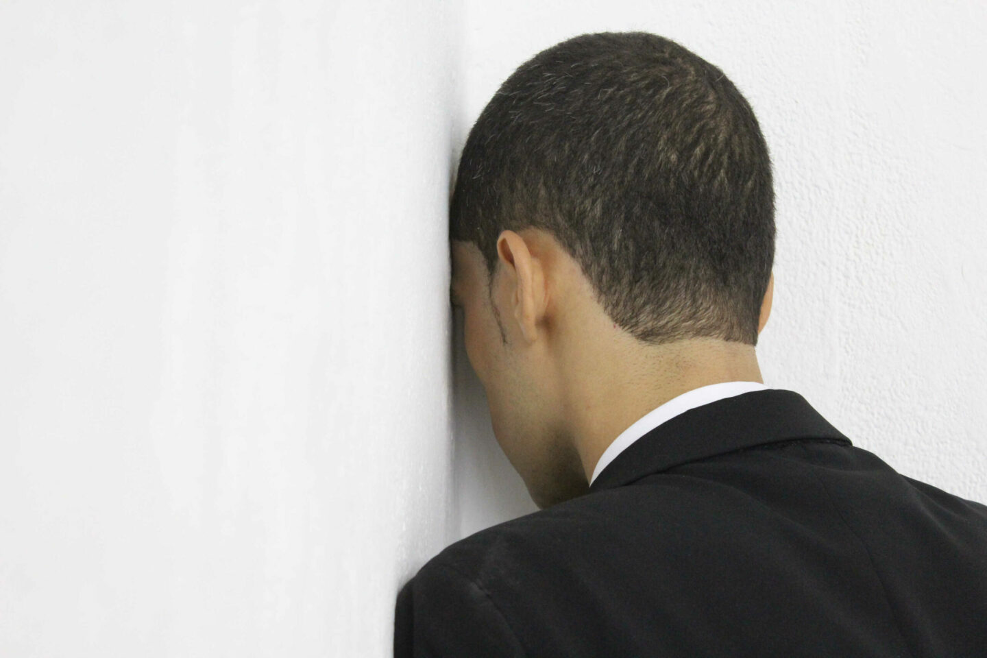 Homem pardo vestido de terno está virado para uma parede, de costas para a câmera, com a cabeça no canto de uma parede.