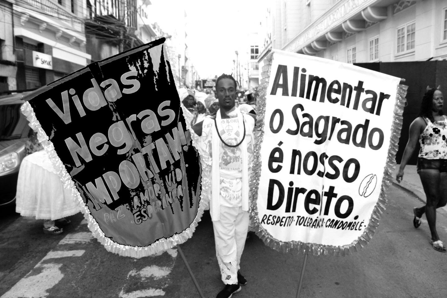 Foto em preto e branco em que um homem negro vestido de branco caminha no meio de uma rua segurando dois estandartes: à esquerda, um deles diz "Vidas Negras Importam" e no lado direito, "Alimentar o Sagrafo é nosso Direito"