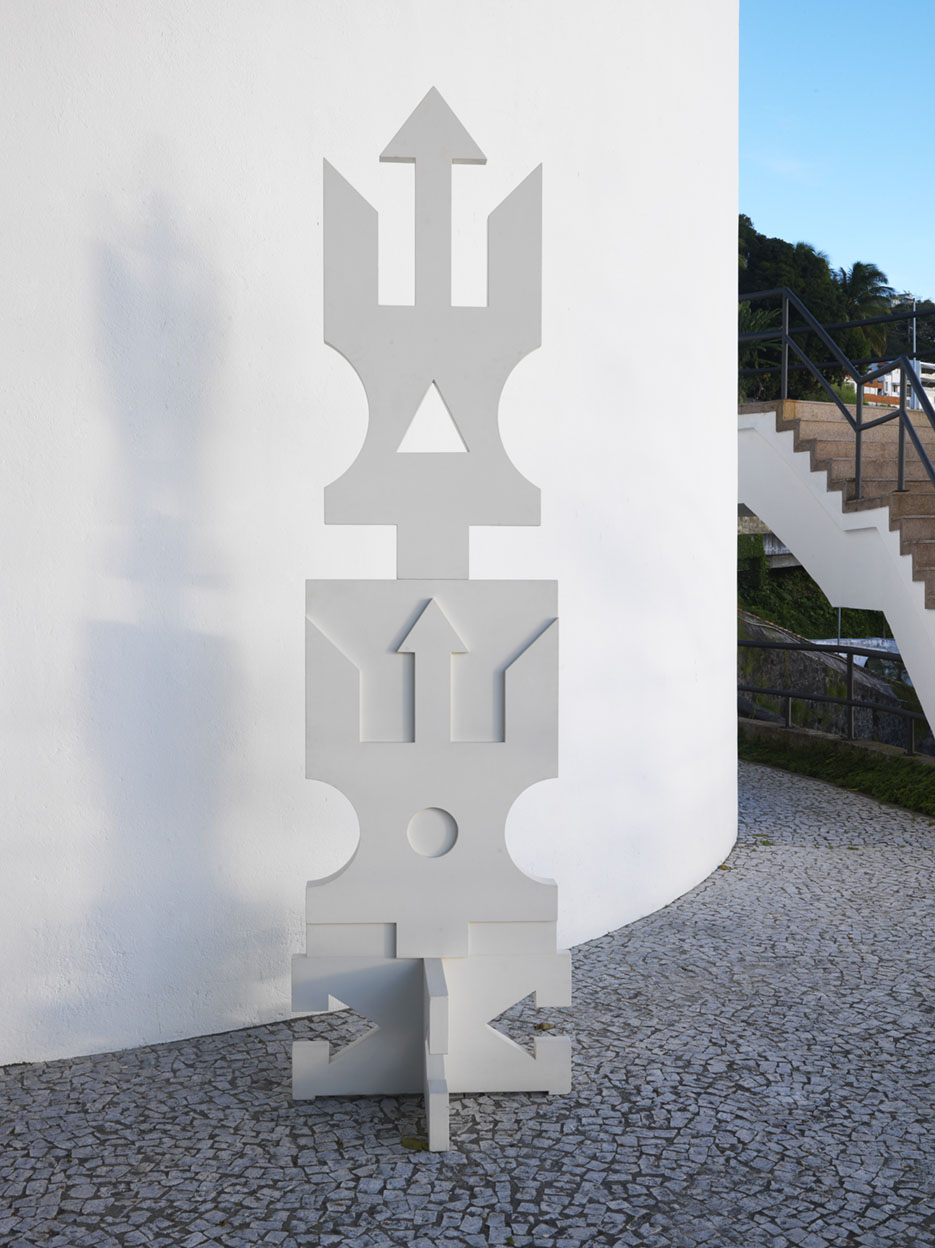 Foto de escultura branca vertical com símbolos afro-brasileiros diante de parede branca.